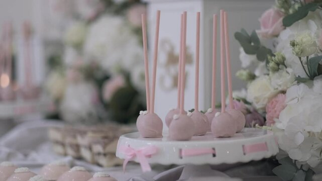 Elegant pink cake pops displayed on a decorated white stand with pastel ribbons, surrounded by delicate floral arrangements, creating a romantic and luxurious dessert table setting