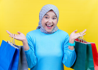 A cheerful asian woman in a hijab with shopping bags enjoys a fun shopping experience against a bright yellow background.