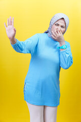 Naklejka premium An asian woman in a hijab and blue outfit holds her nose in disgust, reacting to a bad smell against yellow background.