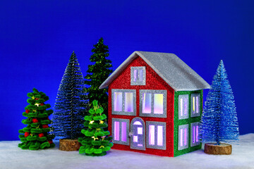 Cozy red green house with soft light in the windows and fir trees around on a blue background