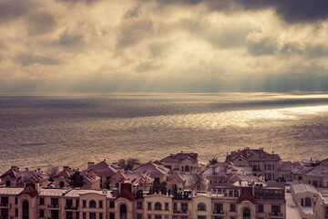 View of houses and rooftops against the backdrop of the sea in cloudy weather, the sun's rays make their way through the dramatic dark clouds over the sea
