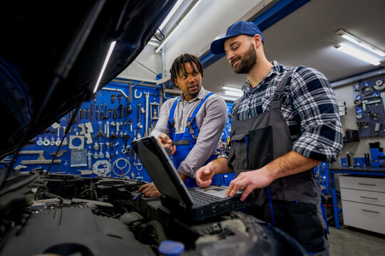 Two auto mechanics are using a laptop computer to diagnose a problem with a car engine in a professional repair shop, showcasing their expertise and modern technology