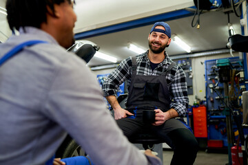 Two cheerful auto mechanics are taking a coffee break and chatting together in their garage,...