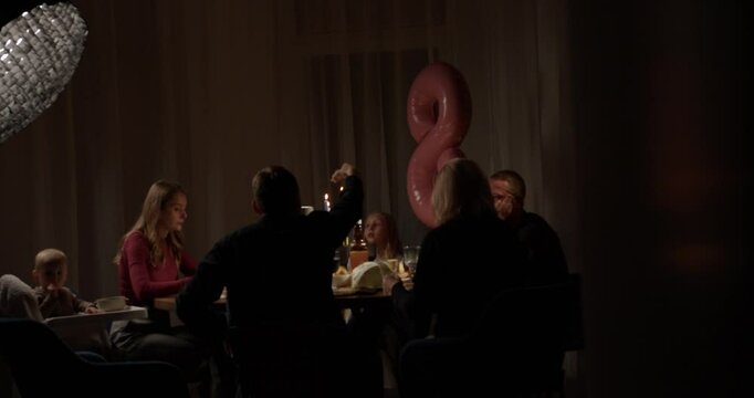 A joyful family gathering takes place with conversations, laughter, and candles illuminating the warm atmosphere.