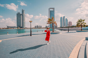 Female tourist wearing red dress walking on promenade in Abu Dhabi and talking on the phone with...