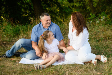 Fototapeta premium Family sitting happily together on cozy blanket spread on green grass. Mom, dad and daughter enjoying time outdoors spent together, talking, laughing. Happy family concept, vacation, family values.