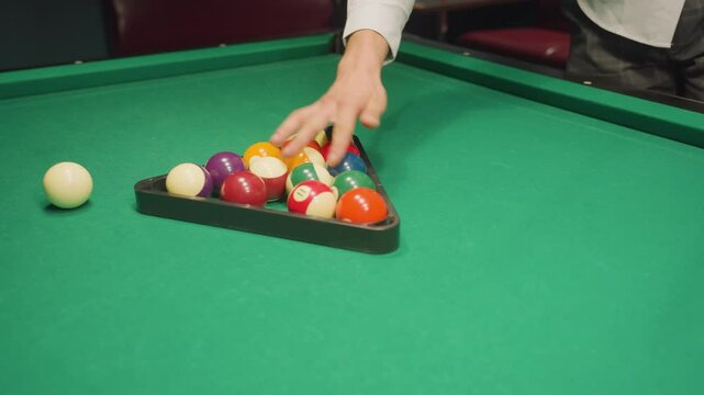 Player hand carefully arranges colorful billiard balls inside triangle rack on green pool table, preparing for break shot in pool hall. Close-up shot captures precise setup before game begins