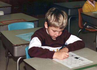 Circa 1987 - Minnesota: Student (boy) in first grade learns to tell time, sitting at his desk in elementary school classroom. Vintage scan, may have imperfections
