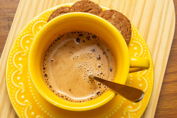 Uma xícara amarela com cappuccino e alguns cookies no píres sobre superfície de madeira.