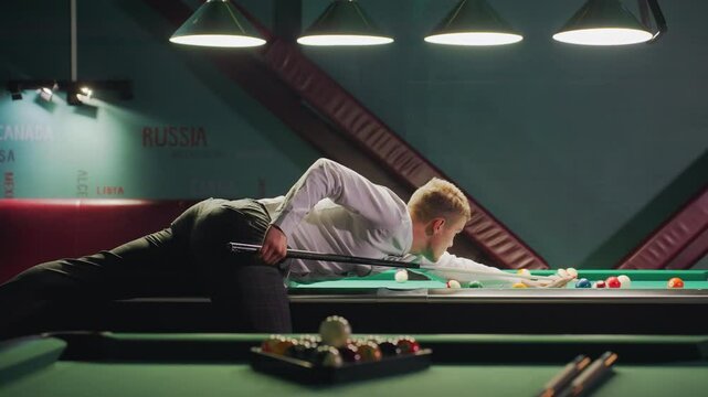 Sportsman in white shirt and grey trousers leans against green pool table, gripping cue stick with intense focus. Dimly lit billiard room enhances concentration as he prepares for precise shot
