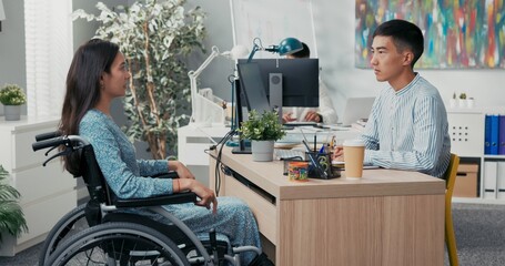 Job interview with disabled woman in wheelchair, stern male boss of Asian-Korean beauty asks questions to girl in dress, they sit in company office on two sides of desk guy listens carefully to answer