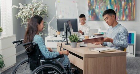 Woman is sitting in wheelchair at desk in office across employer they are signing employee contract...