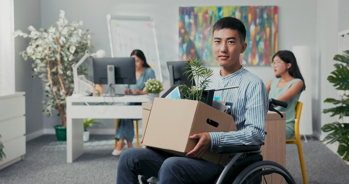 Disabled man dressed in shirt got job in company office he is sitting in wheelchair holding box with essentials for work employee layette looking inside he is smiling happy in background co-workers