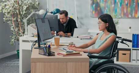 Fototapeta premium Workday in office, corporation, company, co-workers are working on computers, drafting documents, woman in glasses with Asian beauty is sitting at desk in wheelchair, she is disabled, after accident