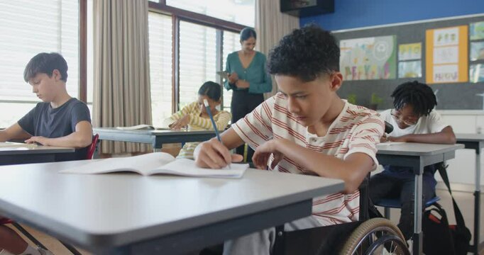 In school, boy in wheelchair writing in notebook, teacher assisting students