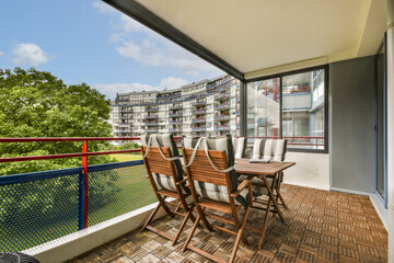 A stylish balcony features wooden chairs and a table, surrounded by a vibrant green tree and contemporary building design in the background.