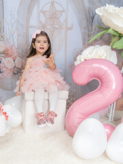 Generated A little girl in pink clothing celebrates in a fairy-tale setting with pastel decorations, a large pink balloon in the shape of a two, and white flowers, radiating joy and innocence.