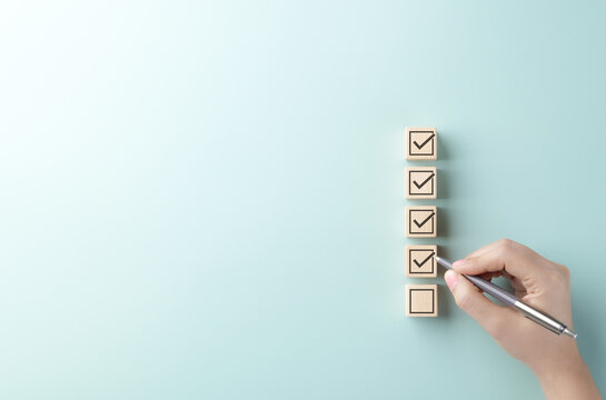 Checklist completion and task management concept. A hand holding a pen marks checkboxes on wooden blocks arranged in a vertical list, symbolizing organization, task management, and goal achievement.