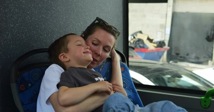 Mother gently tickles laughing son sitting on lap on back passenger seat of bus. Woman makes preschooler kid giggle while entertaining during bus trip