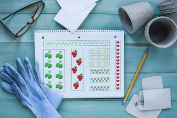 Gardening journal grid notebook with plan surrounded by garden gloves, seeds, glasses, envelopes and peat pots on a rustic wooden table. Table top view.	