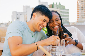 Young Latin man and black woman friends smiling, toasting with beer on a terrace