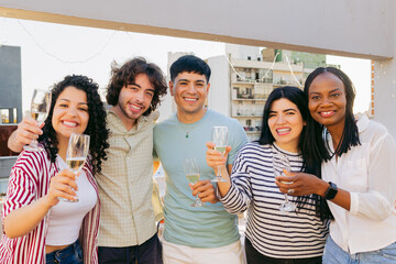Five young Latin friends, men and women, smiling at the camera with a glass of champagne in hand on an outdoor terrace, memory