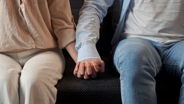 Lovely couple relaxing on the couch at home, close shot of man and woman sit on sofa and touching fingers and holding hands tight.