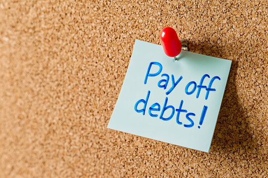 Adhesive note with blue handwriting that says: Pay off debts! It is stuck to a brown cork board with a red push pin.