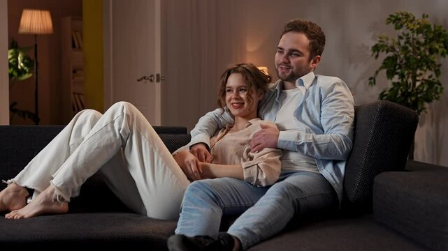 Lovely couple relaxing on the couch at home, man and woman sit in the room watching tv, cuddling and talking, positive happy expression. Slow motion.
