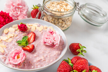Strawberry oatmeal porridge in a bowl with fresh berries and almonds surrounded by fresh strawberries and delicate pink and red roses on white background. Healthy romantic breakfast for holiday