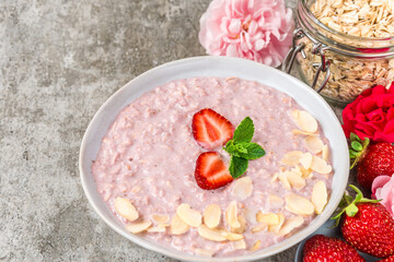 Strawberry oatmeal porridge in a bowl with fresh berries and almonds on concrete background with peony and rose flowers. Healthy romantic breakfast for holiday. Healthy eating and wellness