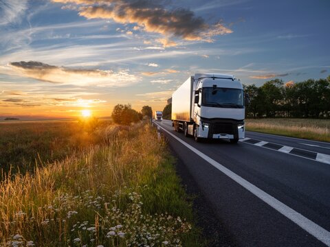 Two white trucks driving on an asphalt road in rural landscape at sunset