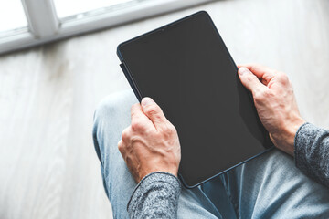 A man holds a black tablet with a blank screen while sitting on the floor near a window, wearing casual jeans and a gray sweater. concept of modern technology and digital lifestyle, copyspace, mockup