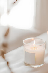 Lit scented candle in a glass jar with a blank label, placed on a soft beige fabric background. The warm glow and neutral tones create a relaxing, minimalist, and luxurious atmosphere