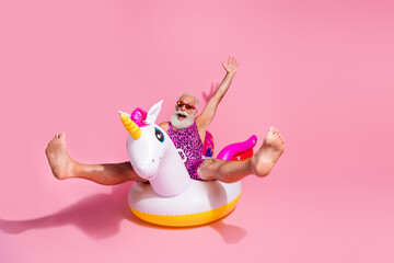 Elderly man in funky leopard print swimsuit on inflatable unicorn with arms up against pink...