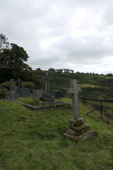 An old English graveyard with weathered tombstones covered in moss, standing amidst lush green grass. Some stones lean slightly, worn by time.