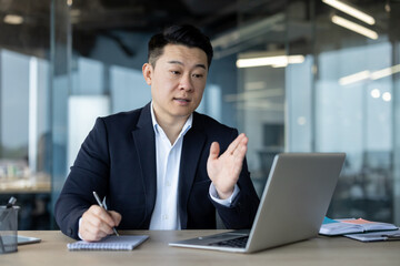 Serious Asian man holding a video conference, talking in the office online on a laptop, explaining, making notes in a notebook