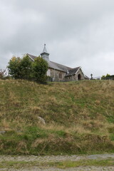 A small, charming stone church sits atop a gentle hill, surrounded by lush green fields in the Welsh countryside. Its weathered walls and modest bell tower stand against a backdrop of rolling hills.