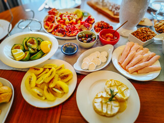 A beautifully arranged breakfast spread featuring sliced kiwi, avocado with lemon, soft cheese, turkey rolls, pomegranate, nuts, and olives for a delicious and nutritious gourmet meal.