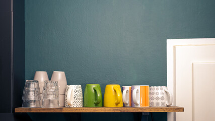 Variety of various cups, mugs and glasses for tea or coffee on wooden shelf in kitchen with colorful wall