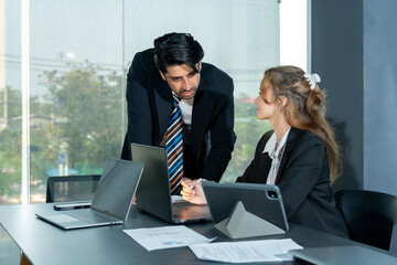 Fototapeta premium Young businesswoman engaging in discussion with businessman leaning forward at modern office desk. Laptops, documents, and tablets arranged on table. Large window showing urban office environment.