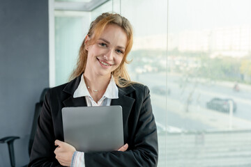 Happy businesswoman holding a laptop in office environment. Confident expression and professional attire indicate success and ambition. Large windows bring natural light into workspace.