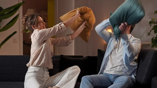 Lovely couple relaxing on couch at home, man and woman in the room playing in pillow fight, laughing joking and hugging, positive expression. Slow motion.