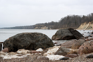 Granite stones and snow remnants on the Baltic Sea coast in spring. In the background is a steep bank and leafless forest. Cloudy sky and fine drizzle creates a haze over the water and the horizon