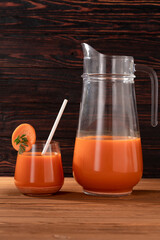 Glass cup with straw and jug with fresh carrot juice. Wooden table under sun rays. Close-up.