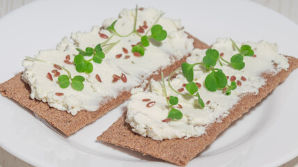 A crispbread with soft cheese, flax seeds, and microgreens on a white plate. A healthy and nutritious snack.