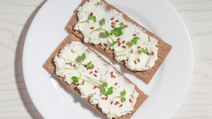 A crispbread with soft cheese, flax seeds, and microgreens on a white plate. A healthy and nutritious snack.