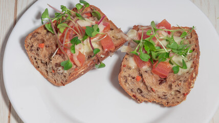 A close-up of two slices of whole grain bread topped with melted cheese, fresh tomatoes, and microgreens, served on a white plate.