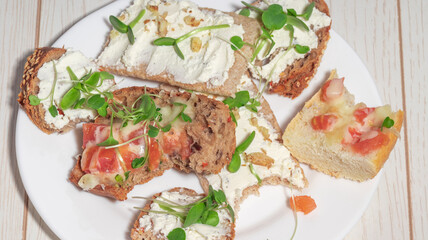 An assortment of open-faced sandwiches with cream cheese, nuts, tomatoes, melted cheese, and microgreens on a white plate.