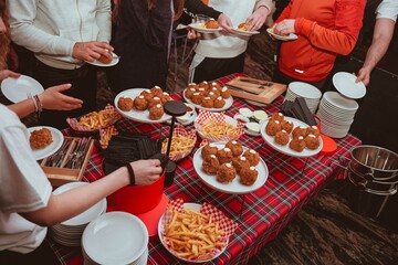 A table set with food during a youth party in a night club. On the table there are plates with supplì filled with mozzarella and baskets of French fries. The people around the table serve themselves.
